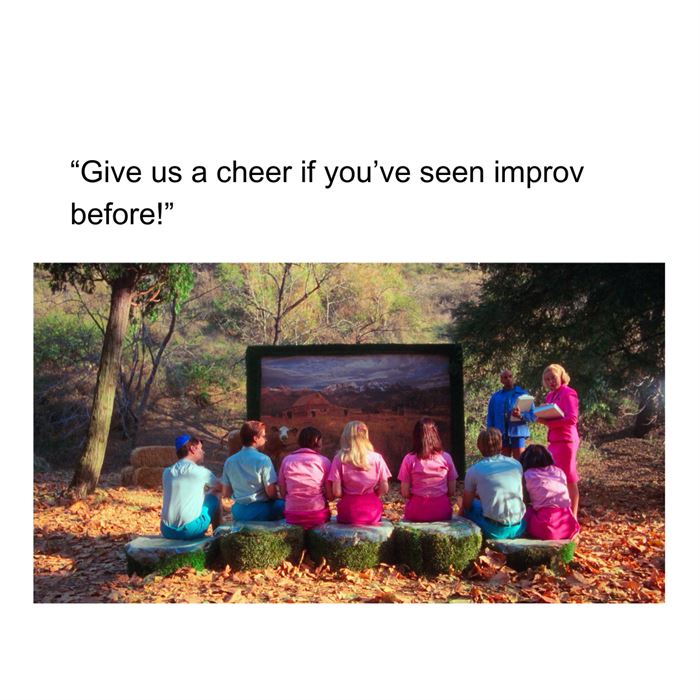 A still from "But I'm a Cheerleader" showing the a group of campers in pink and blue sitting on a log facing a screen with a painted backdrop. There are two counselors facing the group of campers. There is text above saying "Give us a cheer if you've seen improv before!"