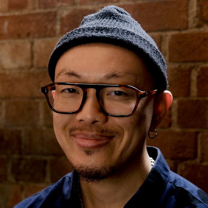 Photo of a Chinese person smiling. He's wearing a black beanie hat, one silver hoop earring can be seen and a dark blue shirt. Behind him is a brick wall.