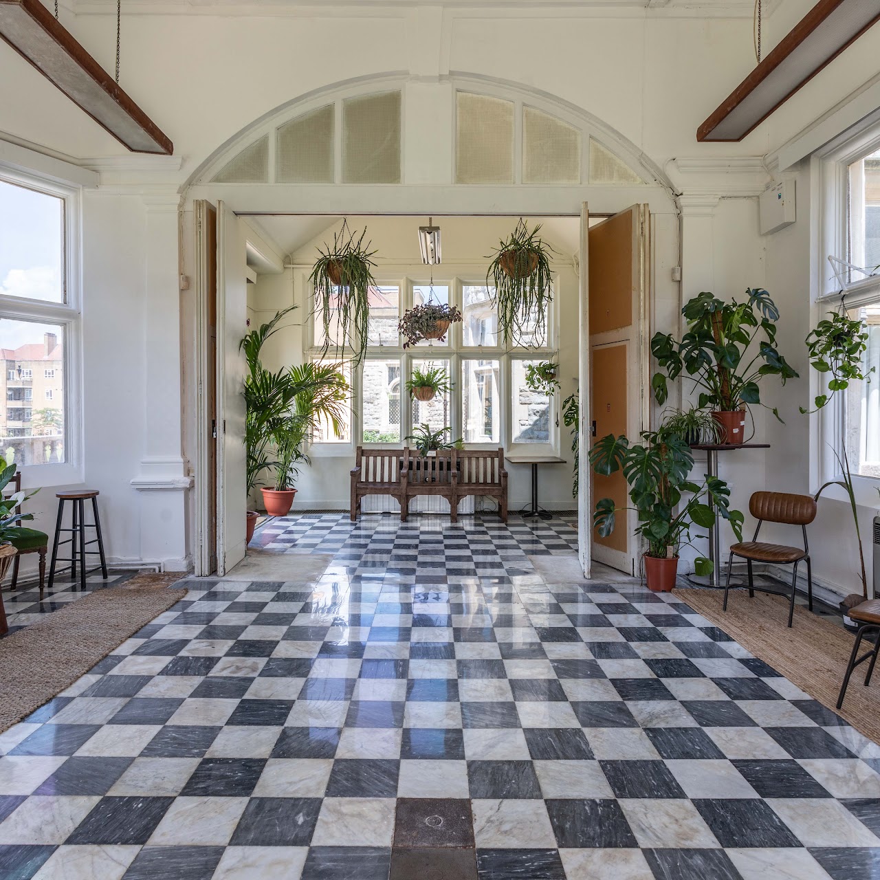 A high ceilinged room with black and white tiled floor and lots of pot plants in the corners and hanging from the ceiling. Big windows let in lots of light, and there are a variety of chairs, benches and stools around the edge of the room