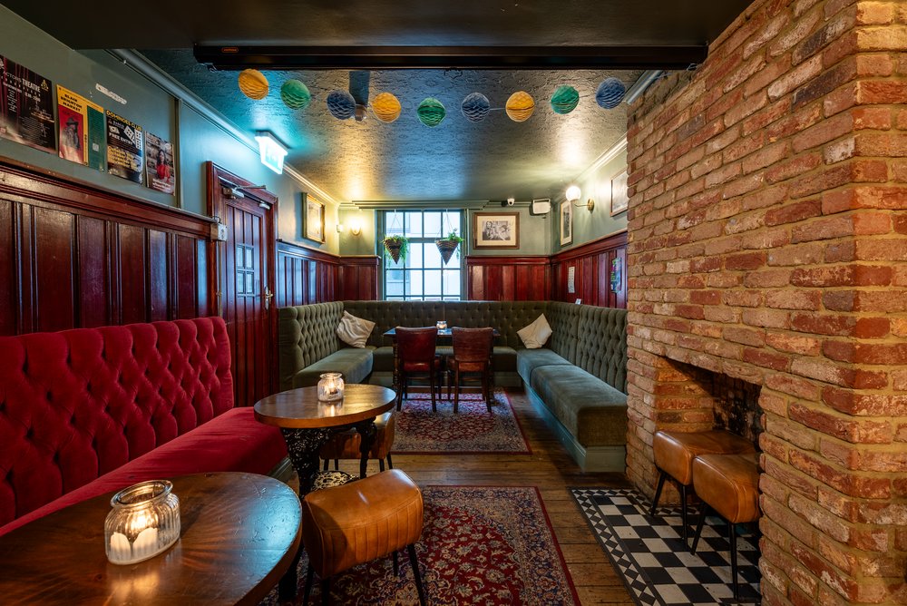 The side-bar at The Actors, a cosy narrow room in an old pub, with wooden panelling, a brick fireplace, patterned rugs on floorboards and candles in holders on the tables. There are posters on the walls and paper decorations hanging from the ceiling.