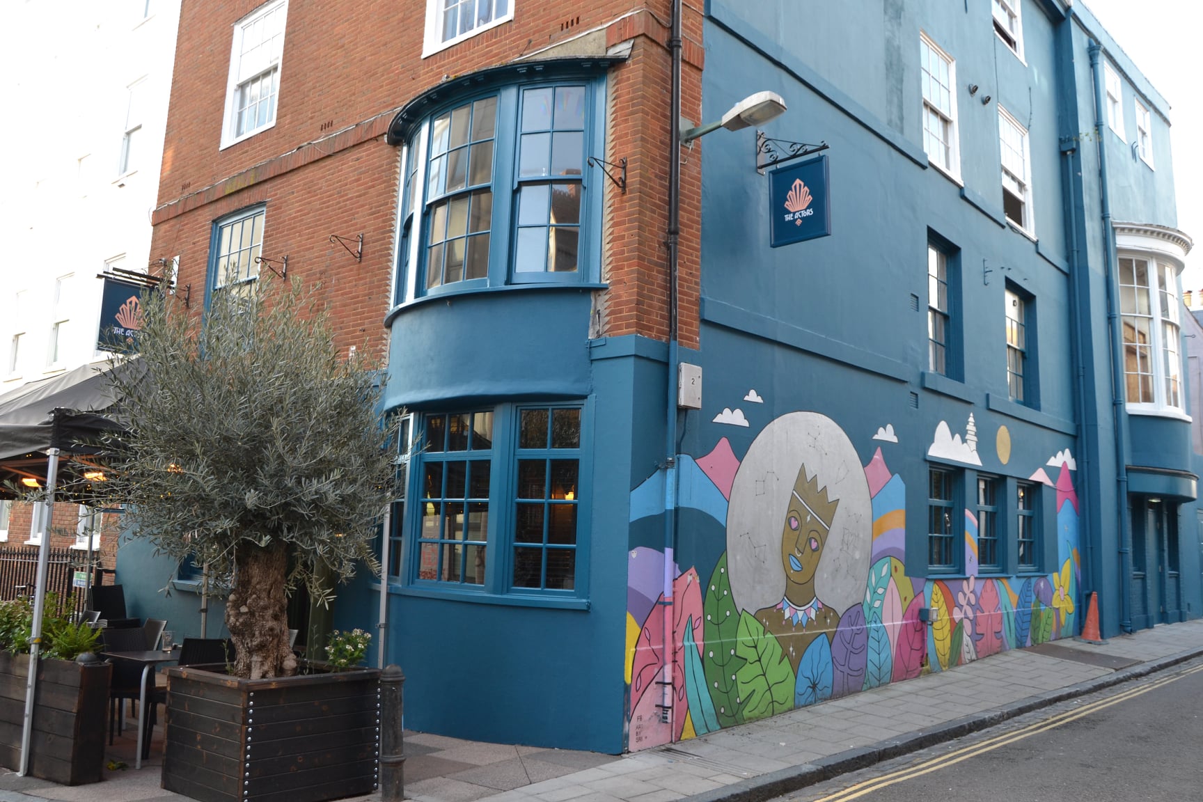 The Actors, an old pub building with curved window bays. Some of the building is red brick, the rest is painted dark teal. There are 2 entrances, one on the left, with plants and tables outside, the other on the right next to a huge painted mural of colourful hills, leaves and a dreamy-looking cartoon person with hearts in their eyes and constellations surrounding their head. 