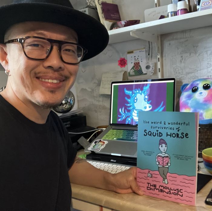 The Mollusc Dimension aka Squid is a light-skinned Asian (Chinese) person wearing glasses, a black hat and holding his comic book smiling. In the background is his desk with a blue dragon on the laptop screen and a rainbow circle bear.