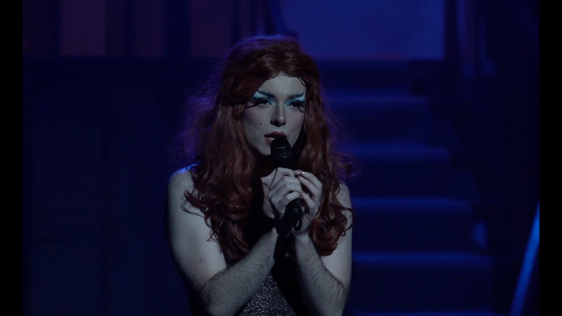 Charlene, a white drag queen in an auburn wig, sings into a microphone on stage. Photograph by El Hakim.